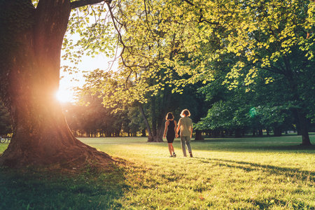 Young long-haired teenage boy with girlfriend walking hand in hand under huge tree during an outdoor walk in a summer city park at sunset time. First love, modern teenagers relations concept image.の写真素材