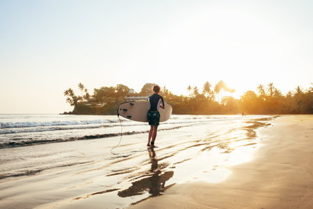 Teen boy with blue and yellow surfboard entering the waves for surfing with sunset rays on palm trees grove. Happy childhood and active sporty people vacation time concept.の写真素材
