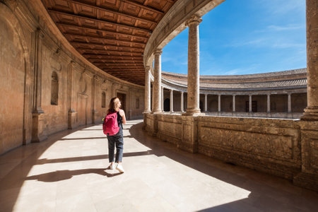 Young female tourist walking with backpack by the wide galleries with columns in the inner circular patio in Palace of Charles V in Alhambra complex in Granada, Andalusia, Spain.のeditorial素材