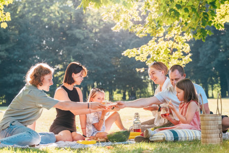 Big family sitting on the picnic blanket in city park during weekend Sunday sunny day. They are smiling, laughing and eating pie with cold tea. Family values and outdoors activities concept.の写真素材