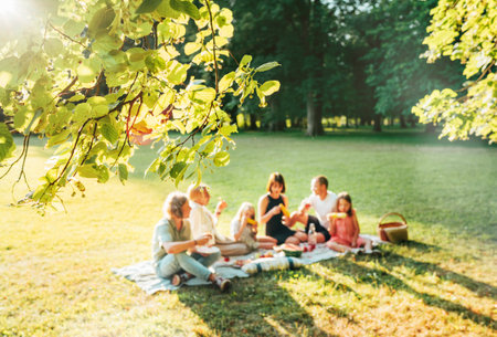 Blossom linden tree branch with Big family sitting on the picnic blanket in city park during weekend Sunday sunny day. They smiling and laughing and eating lunch. Nature outdoors activities concept.の写真素材