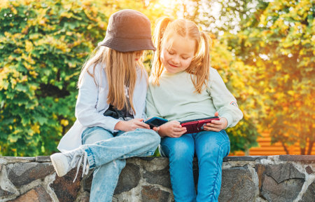 Two smiling little sister girls kids sitting and browsing their smartphone devices in the autumnal park. Careless young childhood time and a modern technology concept image.の写真素材