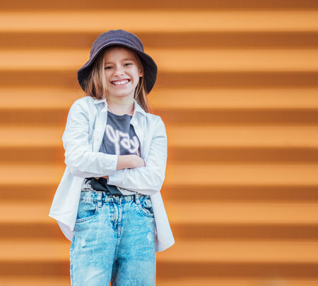 Little fashion-dressed girl portrait sincerely smiling while looking at camera on orange wall background. Urban people living and a street everyday life concept imageの写真素材