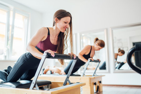 Two young females doing ABS muscles strength exercises using  pilates reformer machine in sport athletic gym hall. Active people training, yoga classes concept.の写真素材