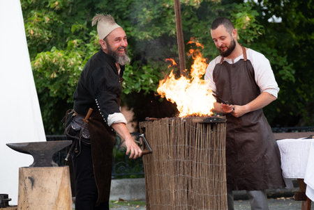 Zilina, Slovak RepubliÑ, Slovakia - July 28, 2023: Two smiling blacksmiths dressed medieval costumes and at anvil during traditional annual restoration known as a "Stredoveky den" in Zilina,のeditorial素材