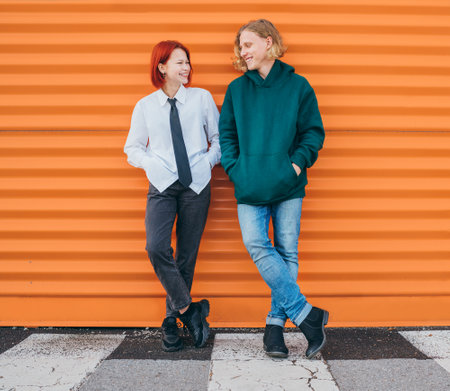 Portraits of two smiling to each other caucasian teen friends boy and girl casual dressed while they standing lean on the orange wall background. Careless young teenhood time and first love concept.の写真素材