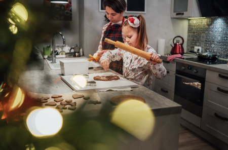 Cute little girl with mother making homemade Christmas gingerbread cookies using rolling pin together in home kitchen.Happy family holidays preparation, childhood through Christmas tree view photo.の写真素材