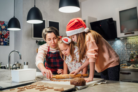 Cute little girls in red Santa hats with mother making homemade dough Christmas gingerbread cookies using cookie cutters together in home kitchen. Happy family holidays preparation, childhood concept.の写真素材