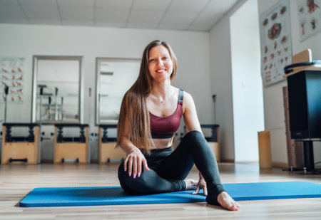 Beautiful fit-shape female dressed sporty sitting and sincerely smiling on sporty gym mat in sport athletic gym. Active people training or medical rehabilitation concept imageの写真素材