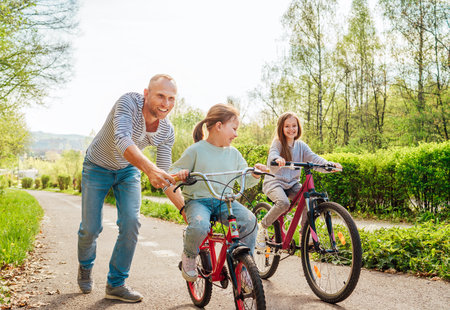Smiling father with two daughters during outdoor walk. He teaching younger girl to ride a bicycle. They enjoy togetherness in the summer city park. Happy childhood concept image.の写真素材