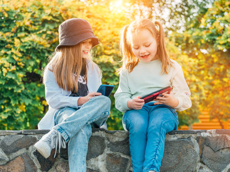 Two smiling little sister girls kids sitting and browsing their smartphone devices in the autumnal park. Careless young childhood time and a modern technology concept image.の写真素材