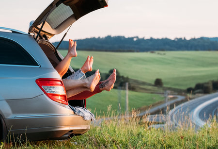 Four-person family lying in the car trunk and fooling around raising feet up. The young caucasian couple has an auto trip break. Family values, traveling, and friendship concept.の写真素材