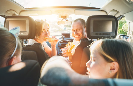 Young couple with daughters eating just cooked Italian pizza sitting in modern car with transparent roof. Happy family moments, childhood, fast food eating or auto journey lunch break concept image.の写真素材