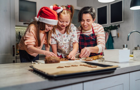 Cute little girls in red Santa hats with mother making homemade dough Christmas gingerbread cookies using cookie cutters together in home kitchen. Happy family holidays preparation, childhood concept.の写真素材