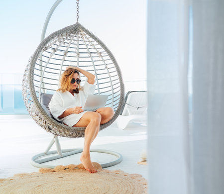 Beautiful middle-aged woman sitting in a comfortable hanging chair on the open house terrace with a modern laptop and browsing in internet doing shopping online. Modern technology and home lifestyle.の写真素材