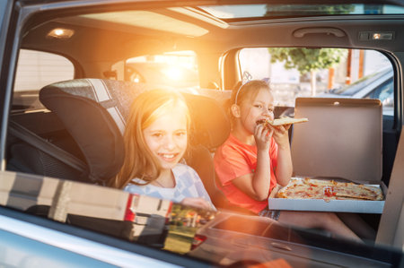 Two positive smiling sisters are happy to eat just cooked Italian pizza sitting in child car seats on the car back seat. Happy childhood, fast food eating, or auto journey lunch break concept image.の写真素材