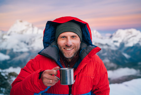 Portrait of smiling at camera high altitude mountaineer dressed red warm dawn jacket holding metal mug of hot tea in with mountains panorama background. Mera Peak High Camp 5700m, Himalayas, Nepal.の写真素材