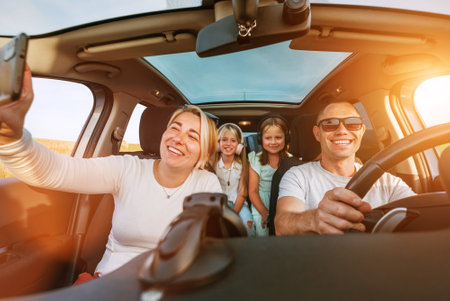 Happy young couple with two daughters inside car during auto trop. They are smiling, laughing and taking selfies using smartphone. Family values, traveling, social media and new technology concepts.の写真素材