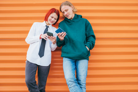 Two caucasian teen friends boy and a girl browsing their smartphone devices while they lean on the orange wall background. Careless young teenhood time and modern technology concept image.の写真素材