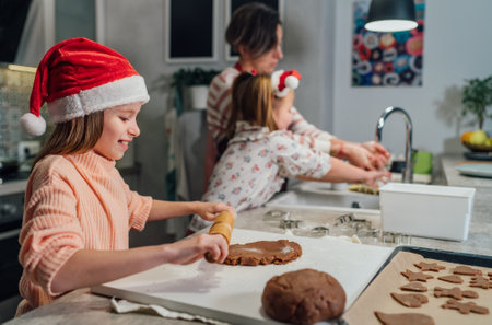 Smiling little girl in red Santa hat kneading Christmas gingerbread dough using rolling pin in home kitchen. Warm family lifestyle moments, happy holidays preparation and childhood conceptの写真素材