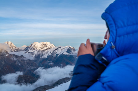 High altitude female mountaineer dressed red warm dawn jacket holding metal mug of hot tea and drinking warm drink with Kangchenjunga mountain panorama. Mera Peak High Camp 5700m, Himalayas, Nepal.の写真素材