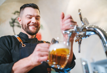 Smiling stylish bearded barman dressed black uniform with an apron tapping fresh lager beer into...