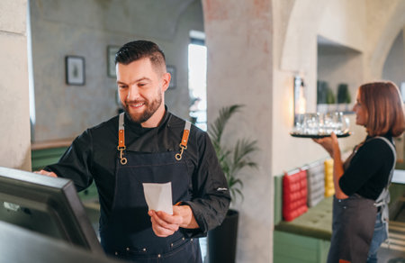 Stylish bearded smiling waiter dressed black uniform using point of sale order terminal system touch Screen and waitress woman with tray. Successful people teamwork, restaurant industry concept image.の写真素材
