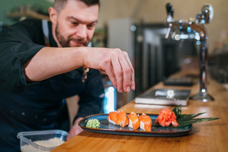 Bearded chef cook dressed black uniform sprinkling the sushi Nigirizushi dish in basement restaurant kitchen. Professional occupation, food preparation, small business and restaurants industry conceptの写真素材