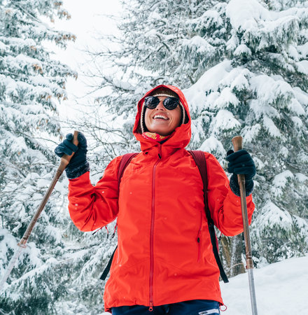 Smiling female trekker dressed red jacket in sunglases with trekking poles walking by snowy slope with fir-trees covered snow, Low Tatra mountains, Slovakia. Beauty in Nature and active people conceptの写真素材