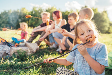 Portrait of little girl eating grilled sausage. Group of Kids - Boys and girls roasting sausages on long sticks over a campfire flame. Outdoor active time spending or camping in Nature concept.の写真素材