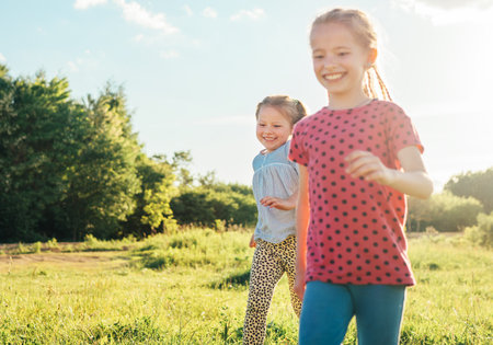 Two little sisters running and laughing in the summer high green grass meadow with a bright sunset light background. Careless childhood, family values, and outdoor activities concept photo.の写真素材