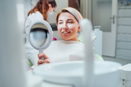 Portrait of young smiling woman sitting in stomatology clinic chair and looked at mirror evaluating her teeth after Tooth whitening procedure. Health care and med care industry concept image.の写真素材
