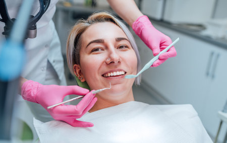 Portrait of young smiling woman sitting in stomatology clinic chair and doctor's hands with dental Mouth mirror and Dental explorer tools preparing to examine teeth condition. Health care concept.の写真素材