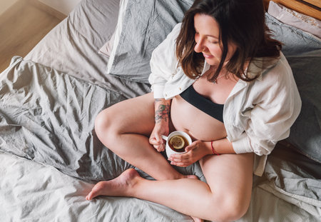 Young pregnant woman sitting with cross-legged on the bed with cup of tea in bedroom in the early morning time. Woman's health, happy pregnancy and calm mental mood concept image.の写真素材