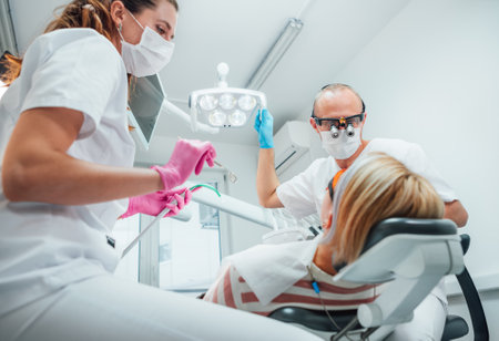 Dental clinic patient appointment. Dentist man in magnifying glasses pointing light. Young female assistant ready for teeth surgery with medical tools. Health care and medicare industry concept image.の写真素材