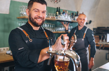 Stylish bearded barman dressed black uniform smiling at camera, beer tapping at bar counter and...