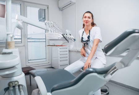 Portraait of sincerely smiling young dentist woman dressed white medical scrubs uniform sitting in modern dental clinic next to stomatology  chair. Health care and medicare industry concept image.の写真素材