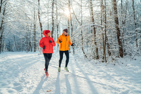 Middle-aged smiling trail couple runners man and woman dressed bright windproof jackets running speaking picturesque snowy forest during sunny frosty day. Sporty active people, winter training image.の写真素材