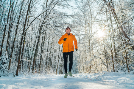 Middle-aged smiling trail runner man dressed bright orange windproof jacket endurance fast running picturesque snowy forest during sunny frosty day. Sporty active people and winter training image.の写真素材