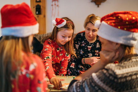 Mother with daughters kids playing together in table board game, wearing Christmas hats. Cozy pre Christmas evening time moment. Family values, board games and X-mas celebration concept.の写真素材