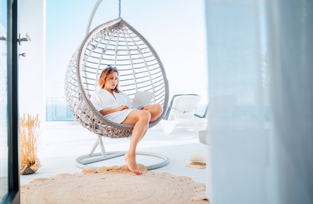 Beautiful middle-aged woman sitting in a comfortable hanging chair on open house terrace with a modern laptop working remotely at home office. Modern technology and distance education and work conceptの写真素材