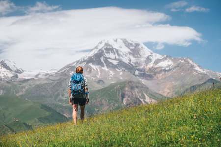 Backpacker woman with a backpack walking and enjoying snowy slopes of Kazbek 5054m mountain while she walking by green grass hill. East Caucasus mountains, Georgiaの写真素材