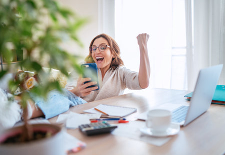 Beautiful middle-aged woman in glasses joyfully laughing at smartphone and making YES gesture at  home office. Small business, successfully investments, bull market or money savings concept image.の写真素材