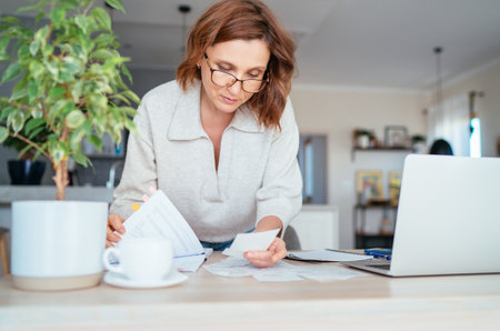 Beautiful middle-aged woman in glasses thinking about family business budget gazing at monthly bills and documents at home living room office. Small business home finances, money savings concept imageの写真素材