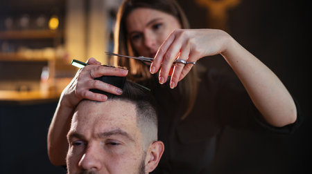 Young hairdresser female hands with scissors and comb hairstyle hairdressing close-up image in modern low light black style barber shop. Haircare service small business Cosmetics and personal careの写真素材