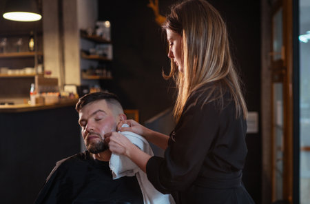 Long-haired young female Barber gently shaving bearded man face bristle while he in chair. She removing cream foam remains from cheeks.Hair and skin care service, local small business concept.の写真素材