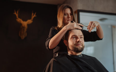 Smiling bearded Man portrait enjoying hairstyle hairdressing in Hair Salon by young hairdresser female. Modern low light black style barber shop interior. Haircare service small business Cosmetics.の写真素材