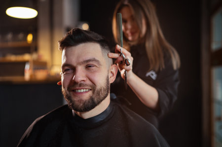 Smiling bearded Man portrait while undercut hairstyle hairdressing in Hair Salon by young hairdresser female. Modern low light black style barber shop interior. Haircare service small business conceptの写真素材
