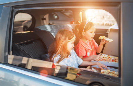 Two little sisters are happy to eat just cooked Italian pizza sitting in child car seats on the car back seat. Happy childhood, fast food eating, or auto journey lunch break concept image.の写真素材