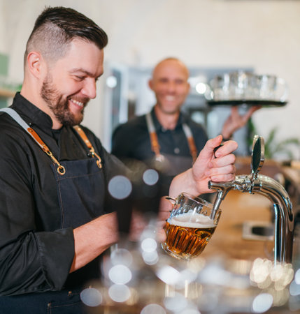 Stylish bearded barman dressed black uniform beer tapping at bar counter and waiter with tray chatting smiling to each other.Successful people team work friendship and good relationships concept imageの写真素材